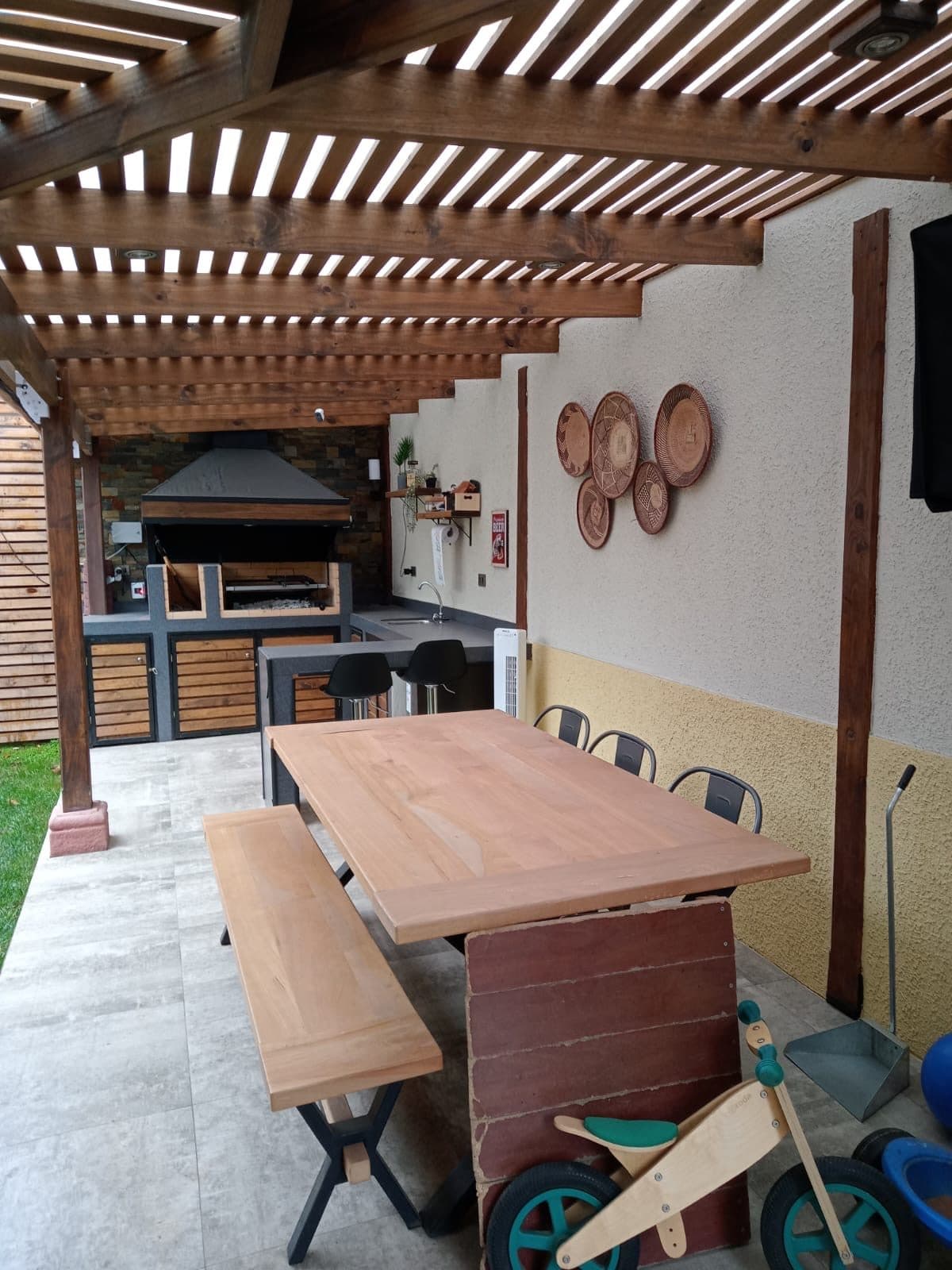 Quincho cubierto por pérgola de madera oscura, con mesa y banca de madera